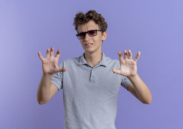 眼镜一个戴着黑眼镜 穿着灰色马球衫的年轻人站在蓝色的墙上做着一个厌恶的防御手势马球站立手势