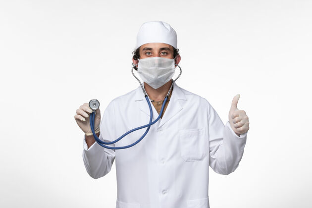 使用正面图：男医生穿着医疗服 戴着口罩以防病毒感染-使用听诊器对白色桌面疾病病毒进行检测-病毒感染-大流行医疗外套冠状病毒预防