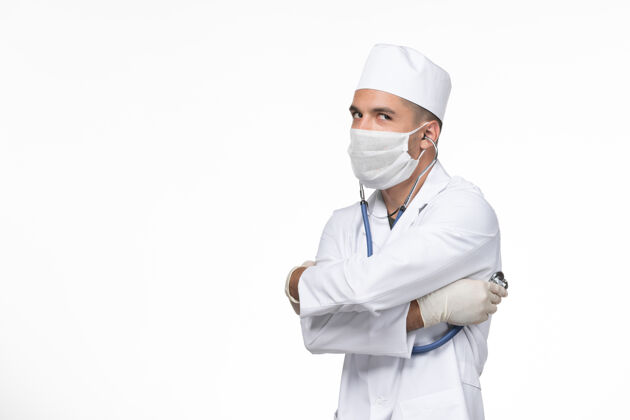 视图正面图：男医生穿着医疗服 戴着口罩 用听诊器对抗冠状病毒 在白墙上传播冠状病毒疾病男性听诊器套装