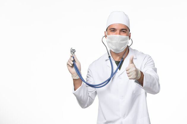听诊器正面图：男医生穿着医疗服 戴着口罩 因为有冠状病毒-用听诊器微笑着对着白墙冠状病毒-大流行性病毒病外套套装口罩