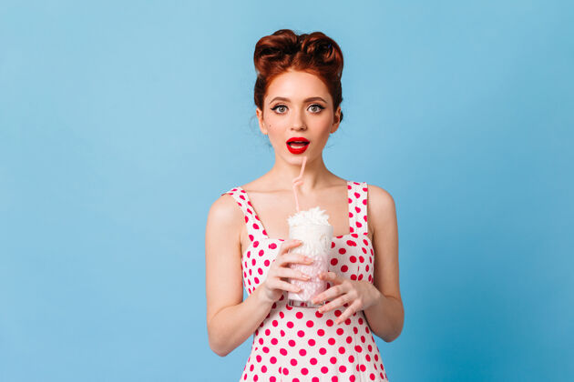 发型惊讶的红唇女孩喝着奶昔摄影棚拍摄的情绪化的年轻女士穿着圆点裙站在蓝色的空间里复古欧式饮料