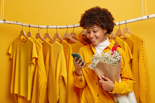 时尚快乐女人第一次约会的照片 站在衣架旁边 在智能手机上收到愉快的短信 手里拿着可爱的花束美国购物消费主义
