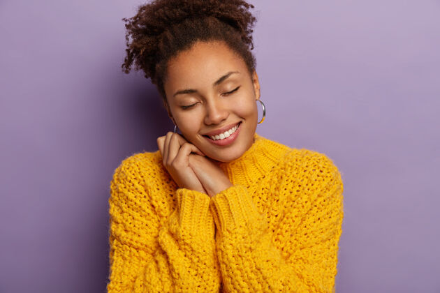 美国快乐的黑皮肤女人 两手都靠着 闭着眼睛 穿着黄色针织毛衣 模特们在紫罗兰色的工作室墙上请休闲编织