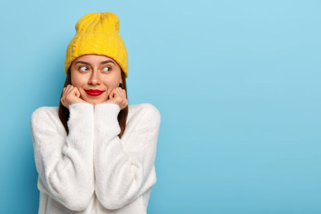毛衣幸福的梦中的欧洲女人的水平镜头穿着最小的化妆品 红色的唇膏 看在一边 穿着黄色的帽子和白色的毛衣 对蓝色背景的姿势 着迷和高兴水平休闲脸庞