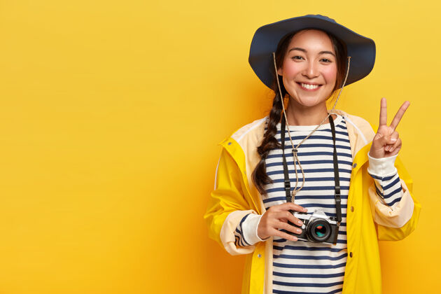 休闲微笑的女旅行者做出和平姿态 用复古相机拍照 戴帽子 条纹毛衣和雨衣 享受刺激的旅行 在黄色背景下摆姿势 复制文本空间深色头发和平自由空间