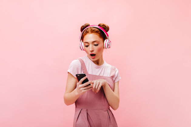 头带穿着粉红色裙子的惊讶的绿眼睛女人的肖像拿着智能手机和耳机听歌曲的女人优雅性感音乐