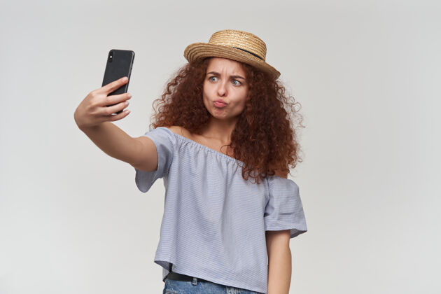 看起来十几岁的女孩 快乐的女人 卷曲的姜黄色头发穿着条纹露肩衬衫和帽子在智能手机上自拍 撅着嘴唇站在白色的墙上孤立模特手机自拍