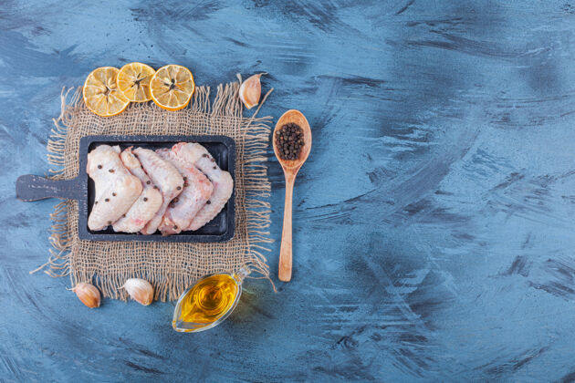 板把鸡翅放在一块粗麻布餐巾的木板上 旁边放着一碗油 香料 汤匙 还有放在蓝色表面的干柠檬香料新鲜配料