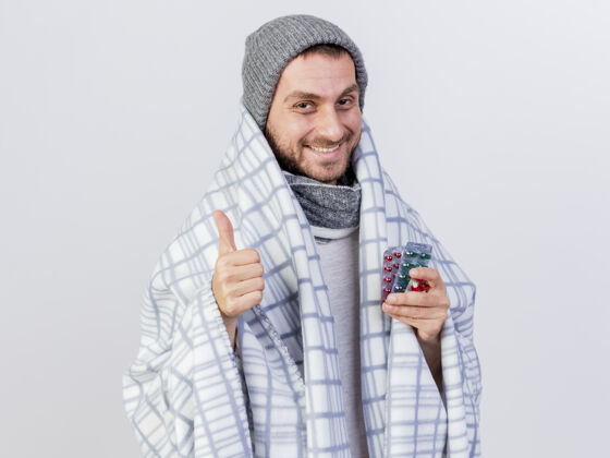 拇指带着冬天帽子和围巾的微笑的生病的年轻人 裹着格纹药片微笑向上抱着