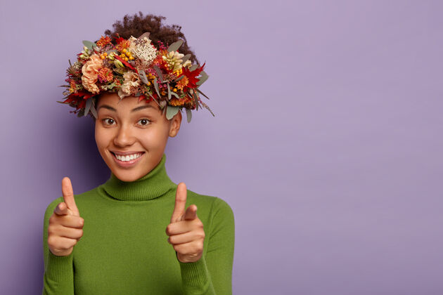 女孩漂亮的美国黑人女孩直指镜头 做手指势 积极微笑 表达自己的选择 戴着自然的秋日植物花环 穿着休闲套头衫 在室内摆姿势手指室内叶子