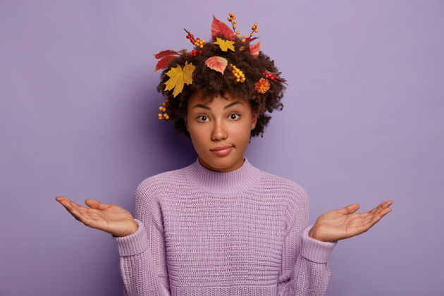 年轻被质问的不知所措的女人摊开手掌 剪着秋天成熟的植物 穿着针织毛衣 不知道看着镜头 孤立在紫色的背景下手势决定树叶