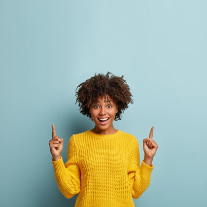 毛衣美丽开朗微笑的女人 非洲式发型 指向上 展示酷的促销或惊人的提议 穿着黄色毛衣 给出建议 在蓝色背景下摆姿势手势肖像手