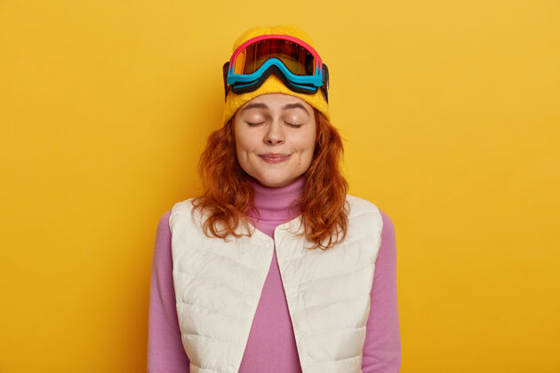 冬天水平拍摄的漂亮年轻女子站在那里 眼睛闭着 穿着温暖舒适的衣服 戴着滑雪面罩滑雪 隔离在黄色背景下黄色滑雪板红发