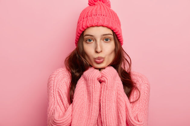 套装美丽的欧洲年轻女子的画像保持嘴唇圆润 手放在下巴下 直视镜头 戴着温暖的冬日帽和毛衣 在粉色背景下摆姿势脸女人站着