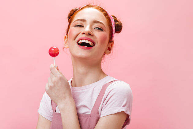 肖像一个穿着白色t恤 有着迷人的粉红色脸颊 笑着拿着棒棒糖 背景是粉色的女人的特写照片年轻时尚红发