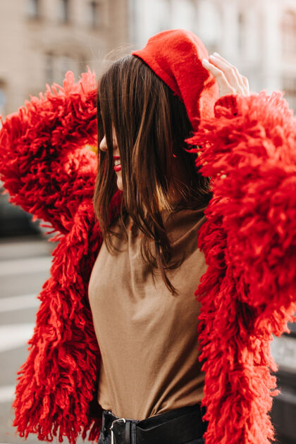 长发正面的直发女人的肖像 穿着蓬松的皮草大衣 t恤和贝雷帽 背景是建筑美丽红色夹克时尚