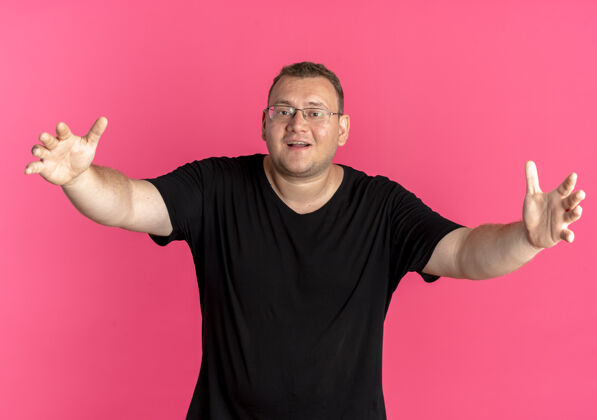 男人戴眼镜的超重男子身穿黑色t恤 双手站在粉红色的墙上做欢迎手势手势手欢迎