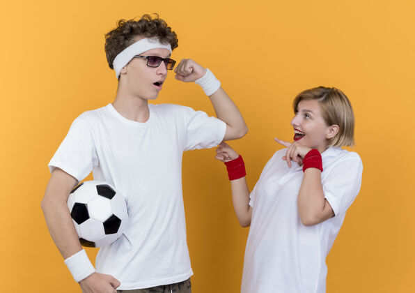 表演一对年轻的运动型情侣站在橙色的墙上 拿着足球向他惊讶的女友展示二头肌运动男人二头肌