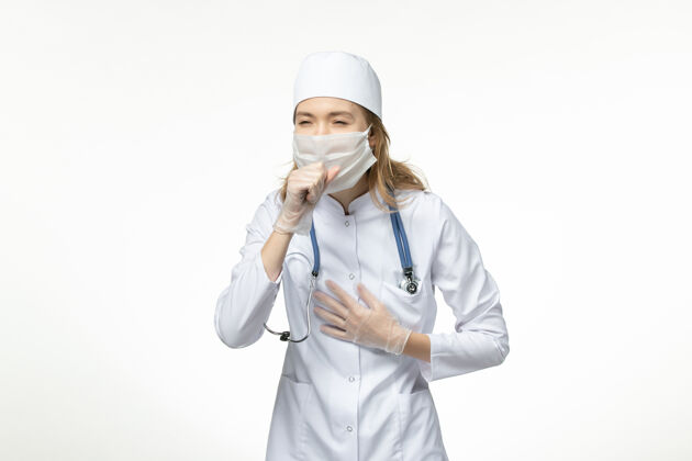 大流行正面图年轻女医生因冠状病毒在白色表面咳嗽 身穿医疗服 戴防护口罩正面到期听诊器