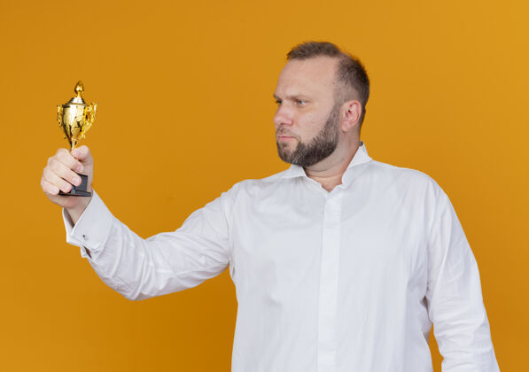 拿着留着胡子的男人穿着白衬衫拿着奖杯严肃地看着它站在橙色的墙上站着奖杯脸