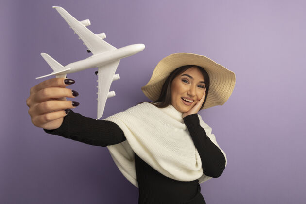 欢呼戴着白色围巾 戴着夏帽的年轻女子展示着玩具飞机欢快的笑容年轻人微笑快乐