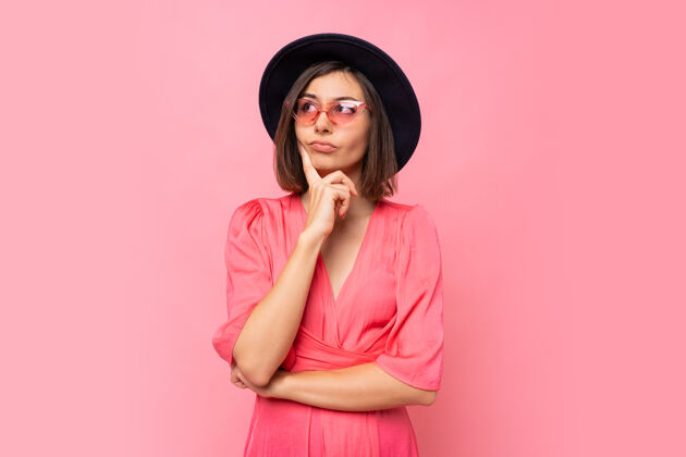 困惑体贴的黑发女人戴着时髦的眼镜在粉红色的墙上摆姿势思考怀疑衣服