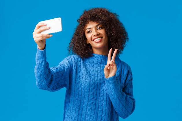手机开朗友好可爱的非洲裔美国女学生自拍在新的智能手机应用程序中应用过滤器 自拍倾头可爱微笑 做和平手势 蓝色墙壁非洲时尚电话