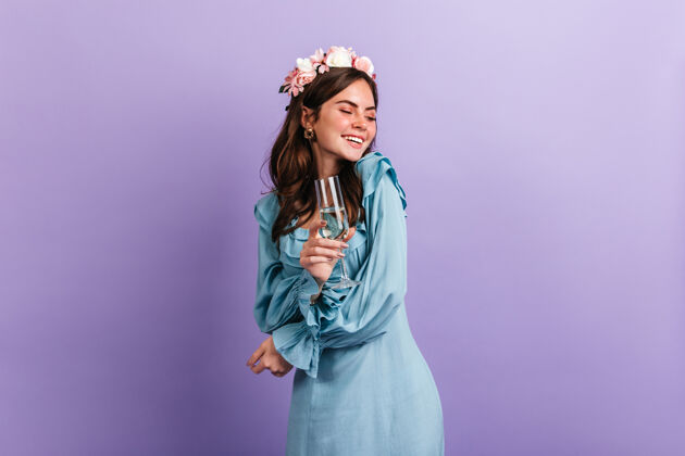 发型积极的女孩兴高采烈地笑着 一边享受着紫色墙壁上的派对身着蓝色服装的模特手里拿着一杯香槟女人白葡萄酒完美