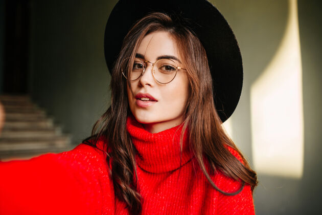 人年轻的欧洲女孩在红色毛衣波浪黑发自拍戴帽子和眼镜的模特摆姿势女人时尚眼镜