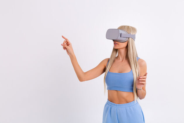 虚拟摄影棚照片中一个年轻迷人的女子穿着温暖的蓝色时尚西装 戴着虚拟现实眼镜在白色的设备快乐表情