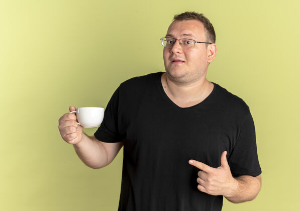 戴着戴眼镜的超重男子身穿黑色t恤 手拿咖啡杯 手指指着咖啡杯 脸上露出幸福的表情 站在明亮的墙上咖啡轻超重