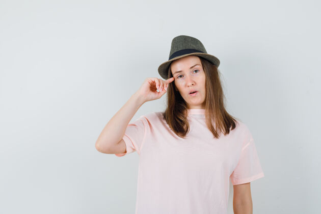 帽子年轻女性手拿手指在太阳穴上穿着粉色t恤 戴着帽子 看起来很懂事前视图年轻敏感女人