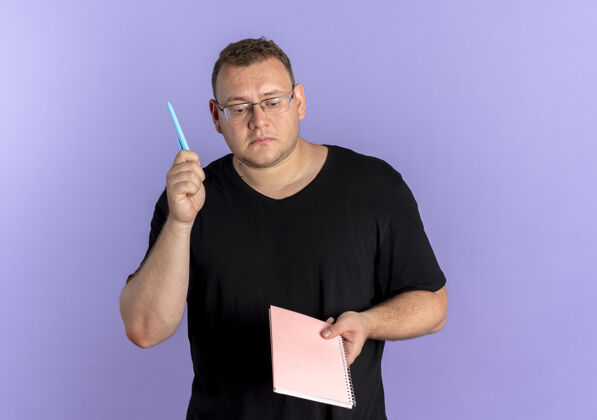 站着一个戴眼镜 身穿黑色t恤 拿着笔记本和钢笔的超重男子站在蓝色的墙上 看上去很困惑钢笔穿看