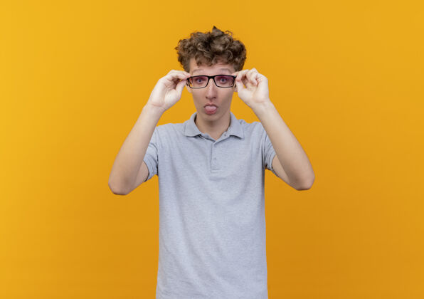 眼镜一个戴着黑眼镜 穿着灰色马球衫的年轻人站在橙色的墙上做鬼脸 伸出舌头马球棍子鬼脸