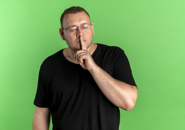 佩戴戴眼镜的超重男子身穿黑色t恤 手指放在嘴唇上 站在绿色的墙上做着沉默的手势手势男人手指