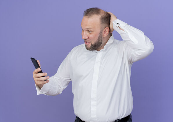移动一个留着胡子的男人穿着白衬衫 站在蓝色的墙上看着他的智能手机 感到困惑看困惑穿