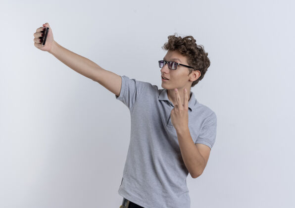 站一个穿着灰色马球衫的年轻人看着他的智能手机屏幕自拍 白色的墙上有一个v型标志看屏幕自拍