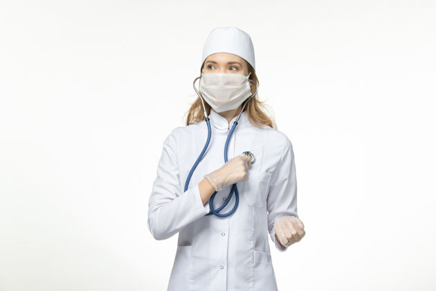 疾病正面图年轻女医生穿着医疗服戴防护面罩因冠状病毒使用听诊器在浅白色表面病毒穿着医生