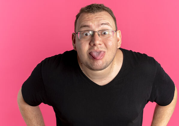 戴着一个戴着眼镜 穿着黑色t恤 伸出舌头站在粉色墙壁上的有趣的超重男人戴着戴着戴着