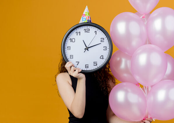 女人一个年轻漂亮的女人 一头卷发 戴着节日帽 手里拿着气球 把脸藏在墙上的挂钟后面 站在橙色的墙上 生日派对的概念脸隐藏卷发