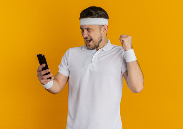 男人身穿白衬衫 头箍金色智能手机 握紧拳头的年轻健身男子站在橙色背景下 快乐而兴奋拳头橙色移动