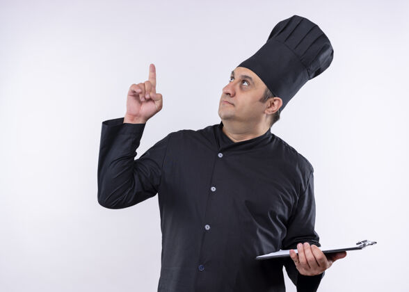 男男厨师身穿黑色制服 头戴厨师帽 手举剪贴板 手指朝上 站在白色背景上黑厨师穿上