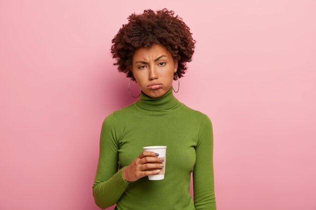 消极不满意疲惫的非洲裔美国妇女拿着外卖咖啡 努力工作后试图提神 穿着绿色的马球领毛衣 疲倦地叹息 感觉工作过度年轻纸发型