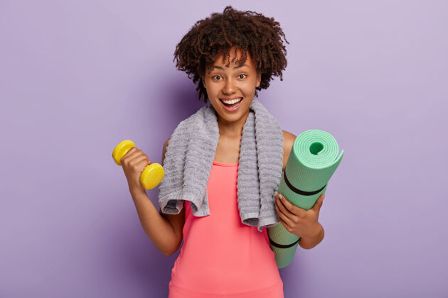活动开朗的黑皮肤非洲年轻女子拿着垫子和哑铃 在健身房锻炼肌肉 有愉快的面部表情 脖子上围着毛巾 穿着粉色上衣 室内模特靠着紫色的墙年轻锻炼黑色