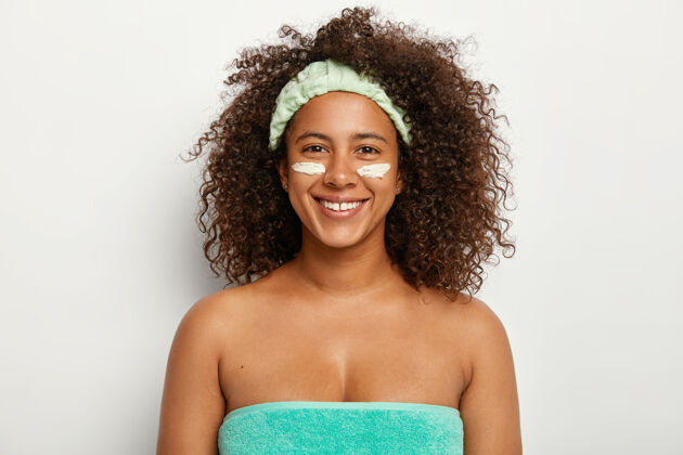 触摸快乐的非洲裔美国女人把面霜放在眼睛下面 皮肤干燥 享受年轻化治疗 裹着毛巾 戴着柔软的头带 积极地微笑女性女士年轻