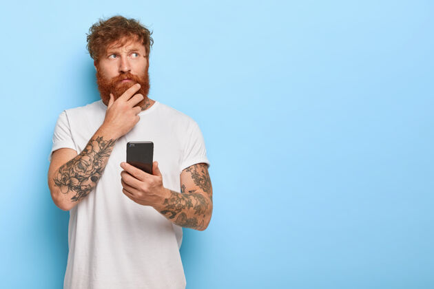 人认真沉思的成年男子横拍摸着浓密的红胡子 拿着手机 上网浏览新闻 想最近的新闻 有纹身的胳膊 穿着休闲的白色t恤男性人类看