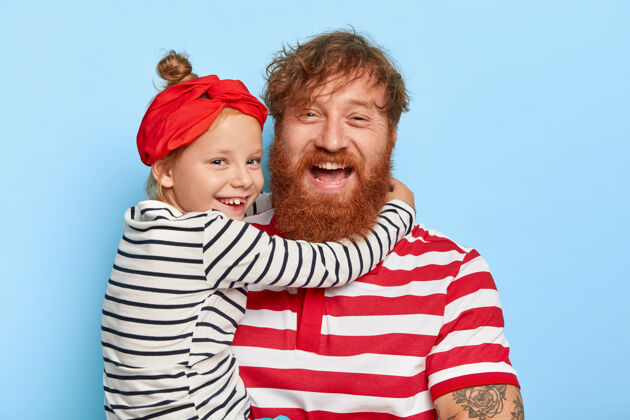 条纹幸福女儿的全家福戴着红色头带和条纹套头衫 用浓密的姜黄色胡须和卷发拥抱着幸福的父亲 彼此非常相爱爱童年满意