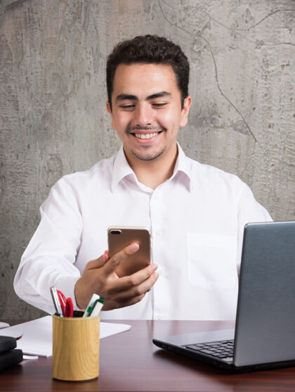 员工微笑的员工看着电话 坐在办公桌旁高品质的照片男办公室笔记本电脑