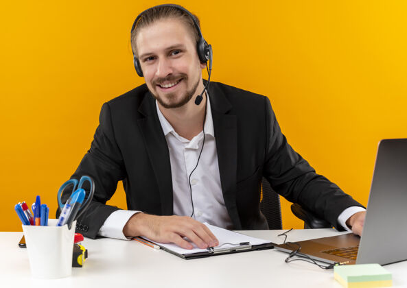 商务人士穿着西装 戴着耳机 拿着麦克风的帅哥坐在橙色背景下的办公桌旁 看着相机微笑着看着笔记本电脑麦克风橙色笔记本电脑