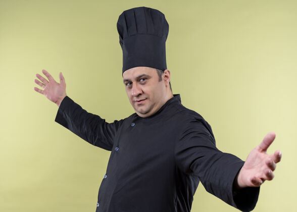 绿色男厨师身穿黑色制服 头戴厨师帽 微笑着做欢迎手势 敞开哈兹站在绿色背景上男性手势欢迎